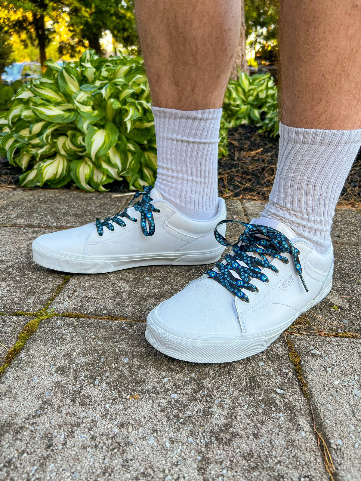 White sneakers with evil eye themed shoelaces worn on a concrete surface with greenery in the background