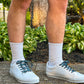 Person wearing white sneakers with evil eye themed shoelaces and beige shorts standing on a concrete surface with greenery in the background.