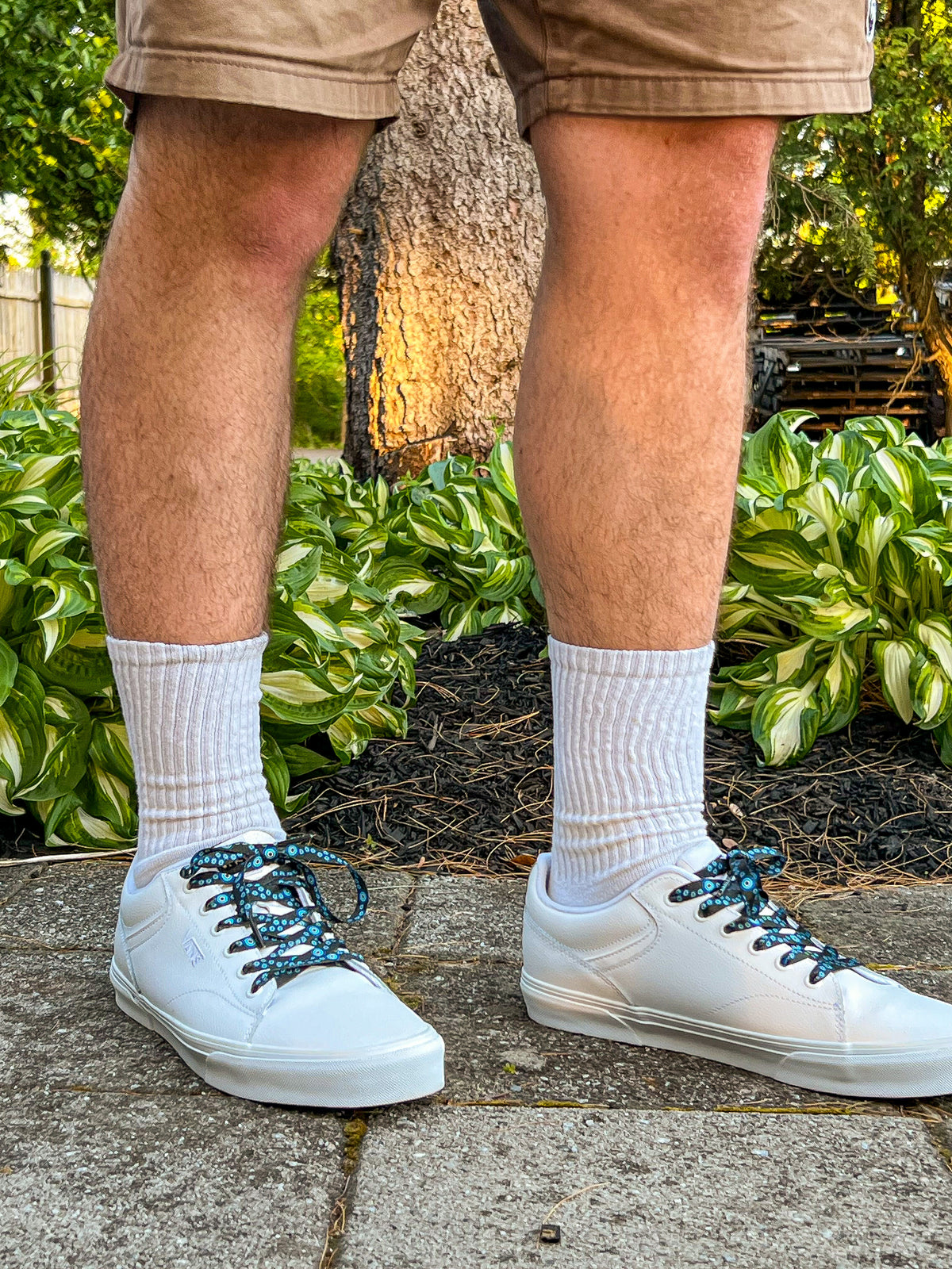Person wearing white sneakers with evil eye themed shoelaces and beige shorts standing on a concrete surface with greenery in the background.