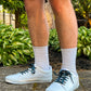 Person wearing white sneakers with evil eye themed shoelaces, white socks, and brown shorts standing on a concrete surface with greenery in the background.