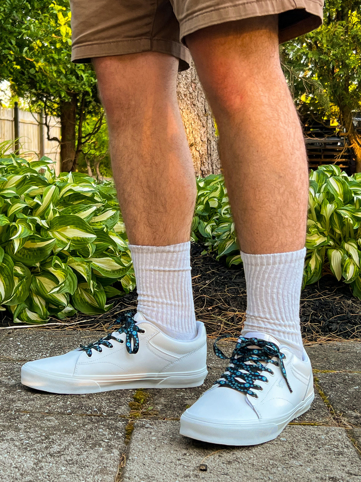 Person wearing white sneakers with evil eye themed shoelaces, white socks, and brown shorts standing on a concrete surface with greenery in the background.