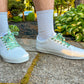White sneakers with green zombie shoelaces worn outdoors on a concrete surface with plants in the background.