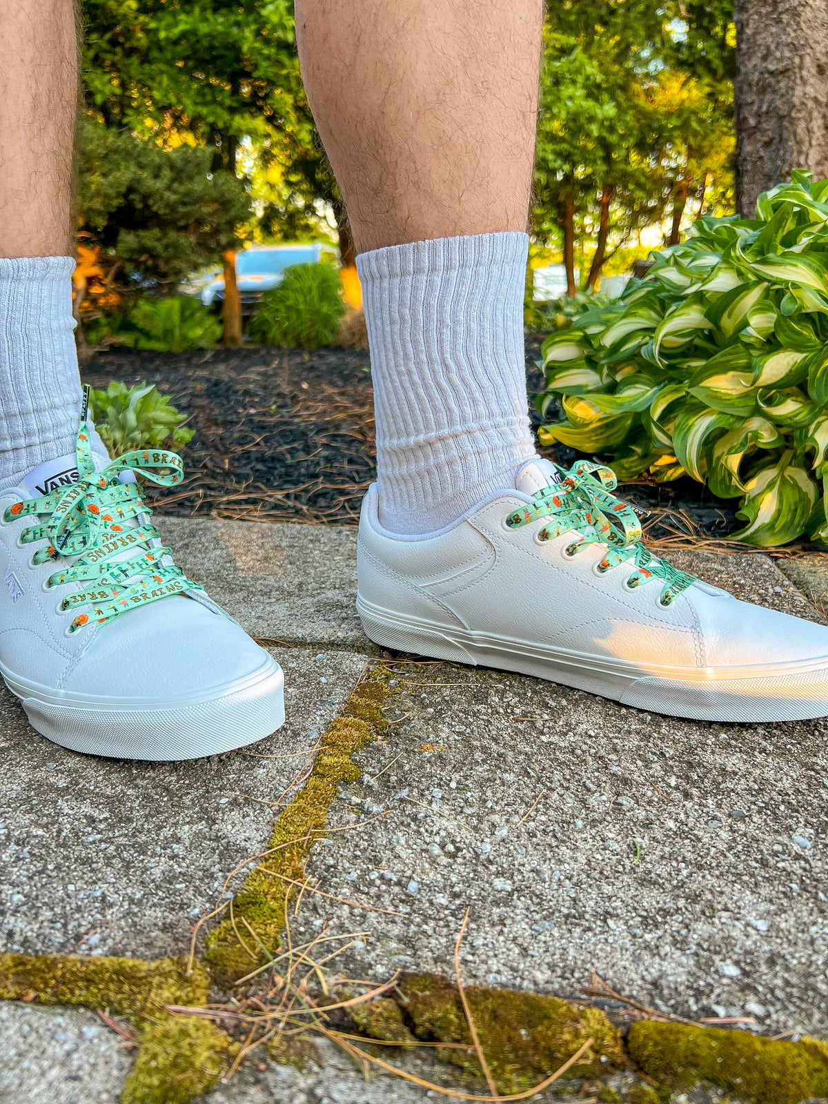 White sneakers with green zombie shoelaces worn outdoors on a concrete surface with plants in the background.