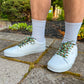 White sneakers with decorative laces worn on a concrete surface with greenery in the background