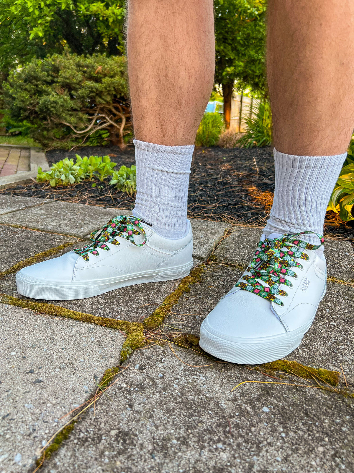 White sneakers with decorative laces worn on a concrete surface with greenery in the background