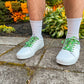 White sneakers with colorful football themed shoelaces worn on a person standing on a concrete surface with plants in the background.