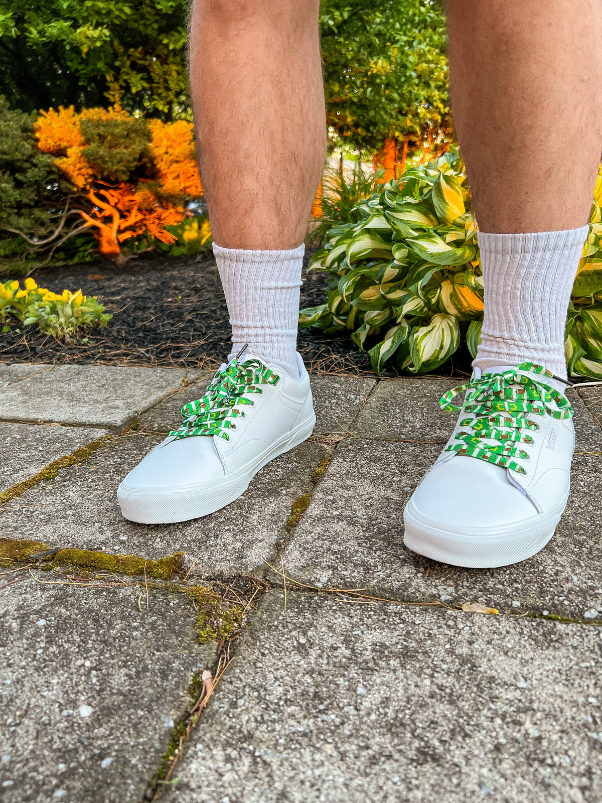 White sneakers with colorful football themed shoelaces worn on a person standing on a concrete surface with plants in the background.