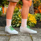 Person wearing white sneakers with green accents, beige shorts, and colorful football themed shoelaces on a concrete surface with plants in the background.