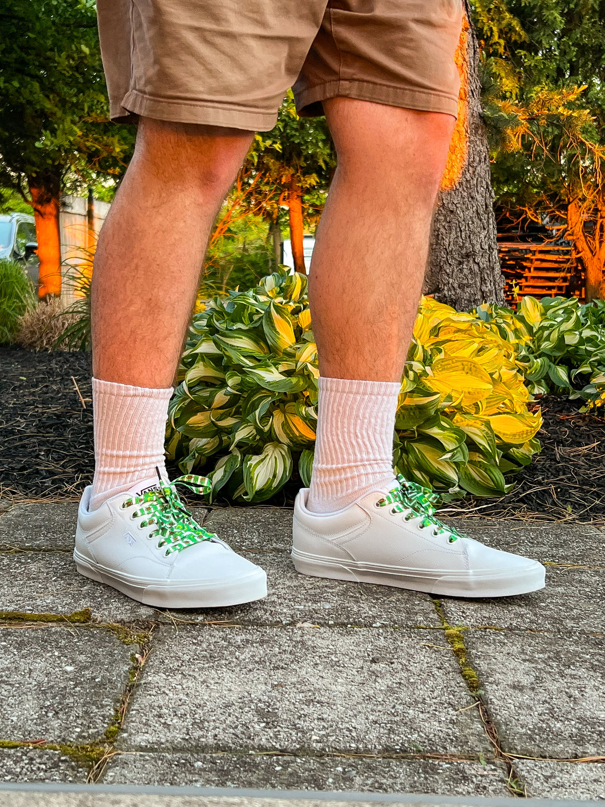 Person wearing white sneakers with green accents, beige shorts, and colorful football themed shoelaces on a concrete surface with plants in the background.