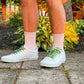 Person wearing white sneakers with colorful football themed shoelaces, pink socks, and brown shorts.