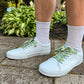 White sneakers with colorful palm tree shoelaces worn outdoors on a concrete surface with greenery in the background.