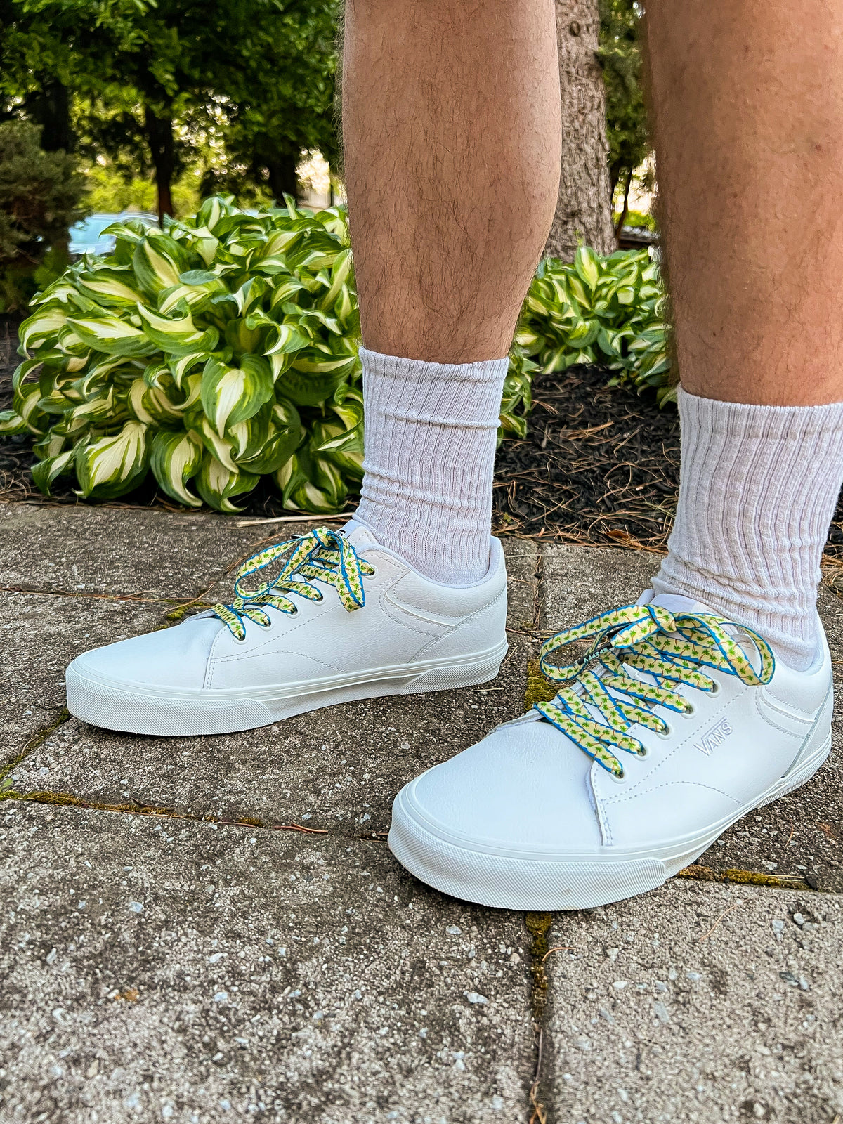 White sneakers with colorful palm tree shoelaces worn outdoors on a concrete surface with greenery in the background.