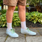 Person wearing white sneakers with colorful palm tree shoelaces standing on a paved area with green plants in the background.