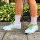 Person wearing brown shorts with  sneakers and colorful palm tree shoelaces on a paved walkway with greenery.