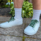 White sneakers with cow shoelaces worn on a concrete surface with plants in the background
