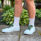 Person wearing white sneakers with cow shoelaces  on a concrete surface with plants in the background