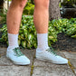 Person wearing white sneakers with cow shoelaces on a stone path with plants in the background
