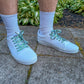 White sneakers with decorative laces worn on a concrete surface with greenery in the background