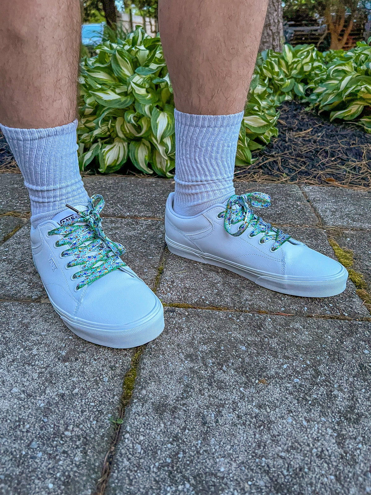 White sneakers with decorative laces worn on a concrete surface with greenery in the background