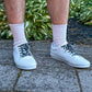 White sneakers with diamond patterned shoelaces worn on a stone path with greenery in the background