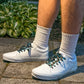 White sneakers with diamond patterned shoelaces worn on a stone surface with greenery in the background