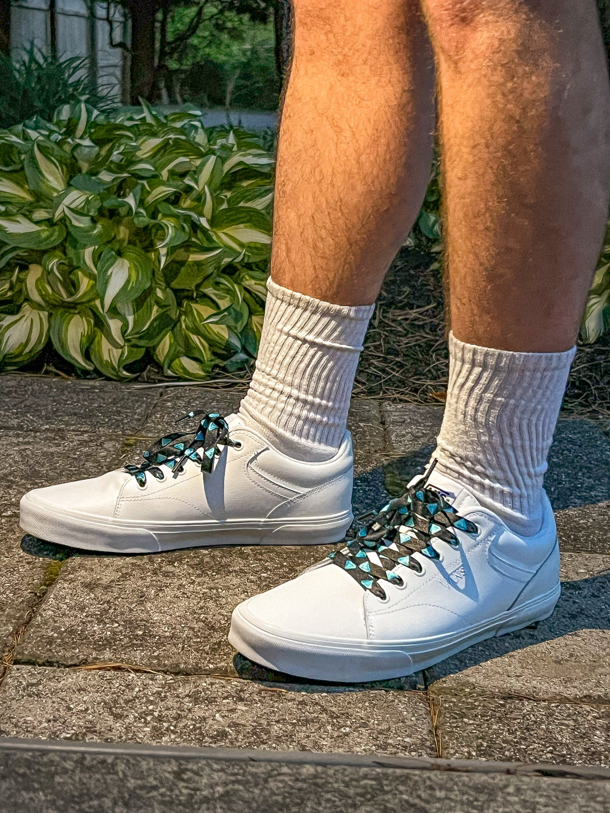White sneakers with diamond patterned shoelaces worn on a stone surface with greenery in the background
