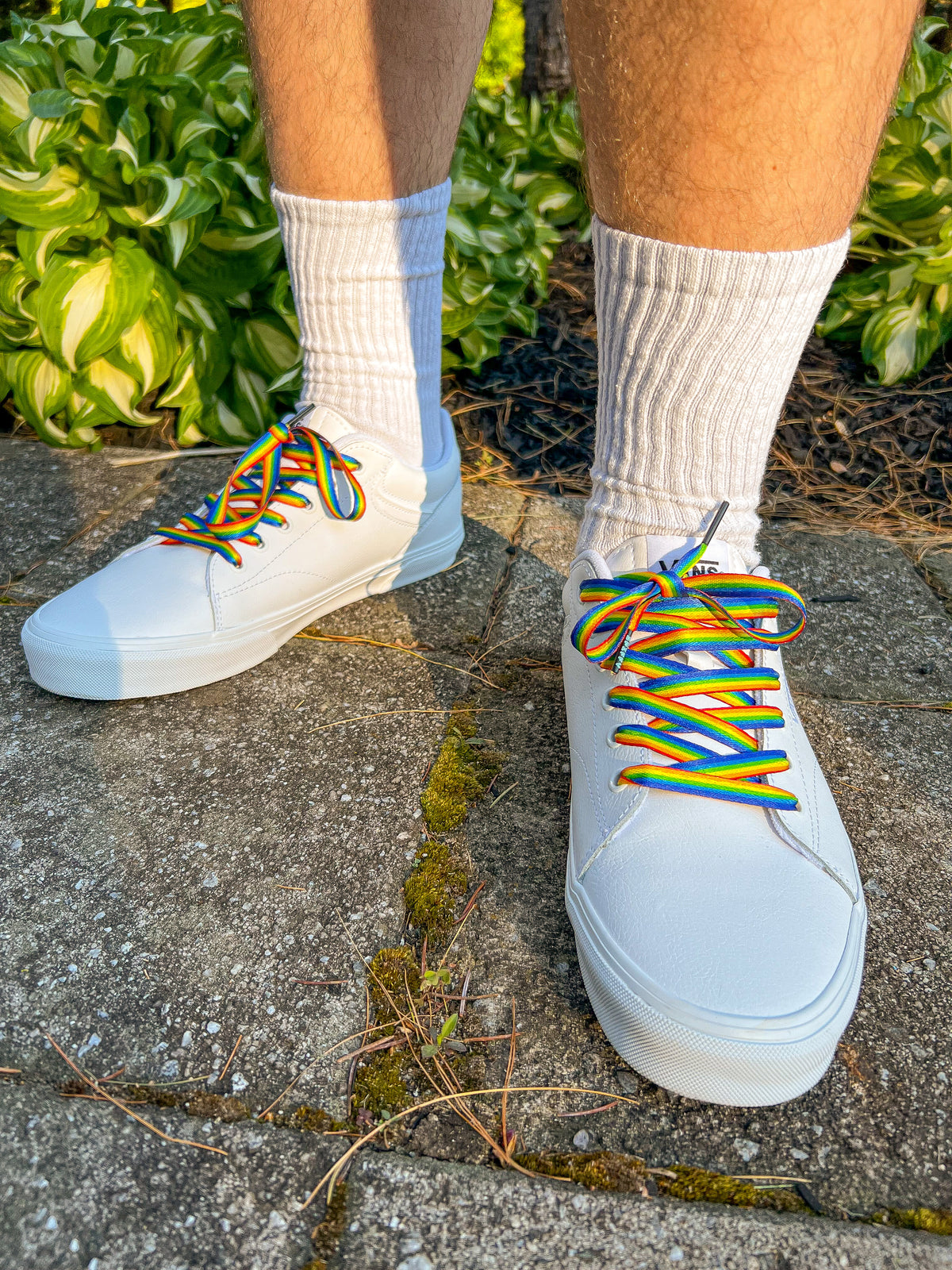 White sneakers with rainbow laces worn on a concrete surface with greenery in the background